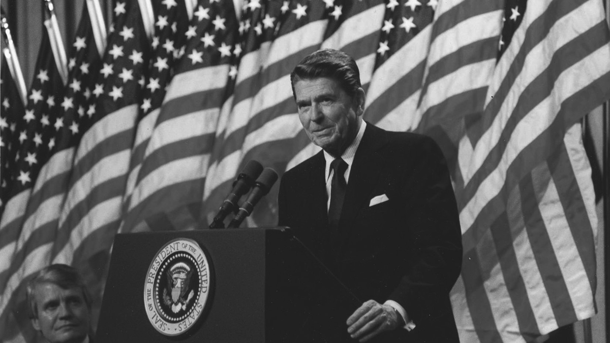 An old man in a suit stands before a phalanx of American flags.