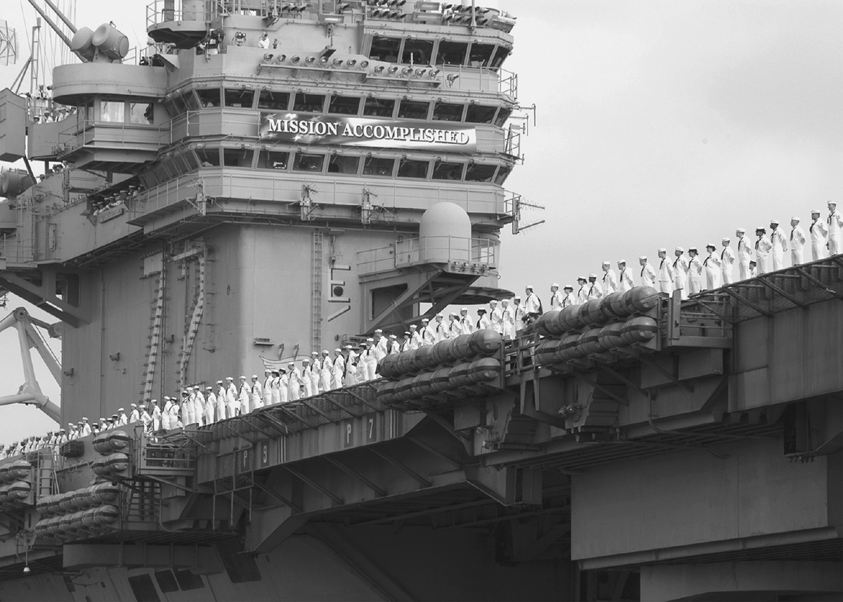 Sailors line the deck of an aircraft carrier with a large banner reading â€œMission Accomplished.â€