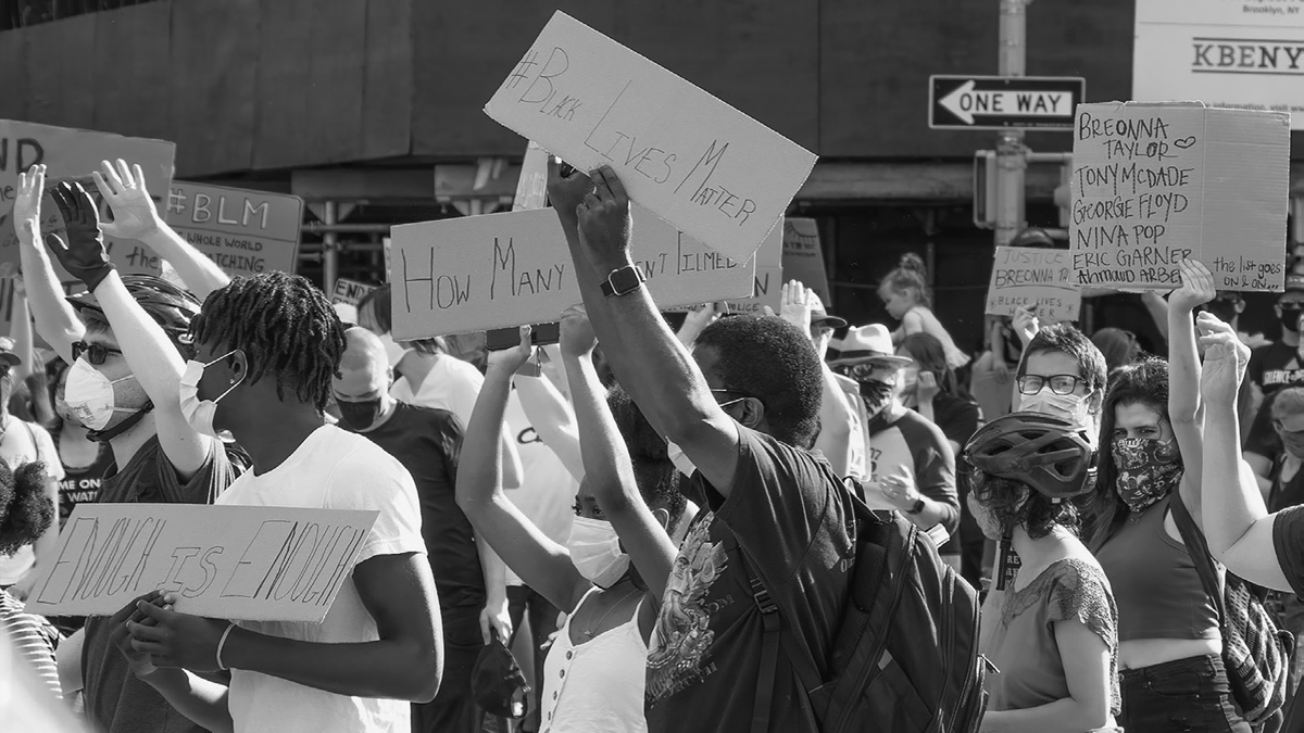 A crowd, wearing medical masks, holds homemade cardboard signs on a street corner.