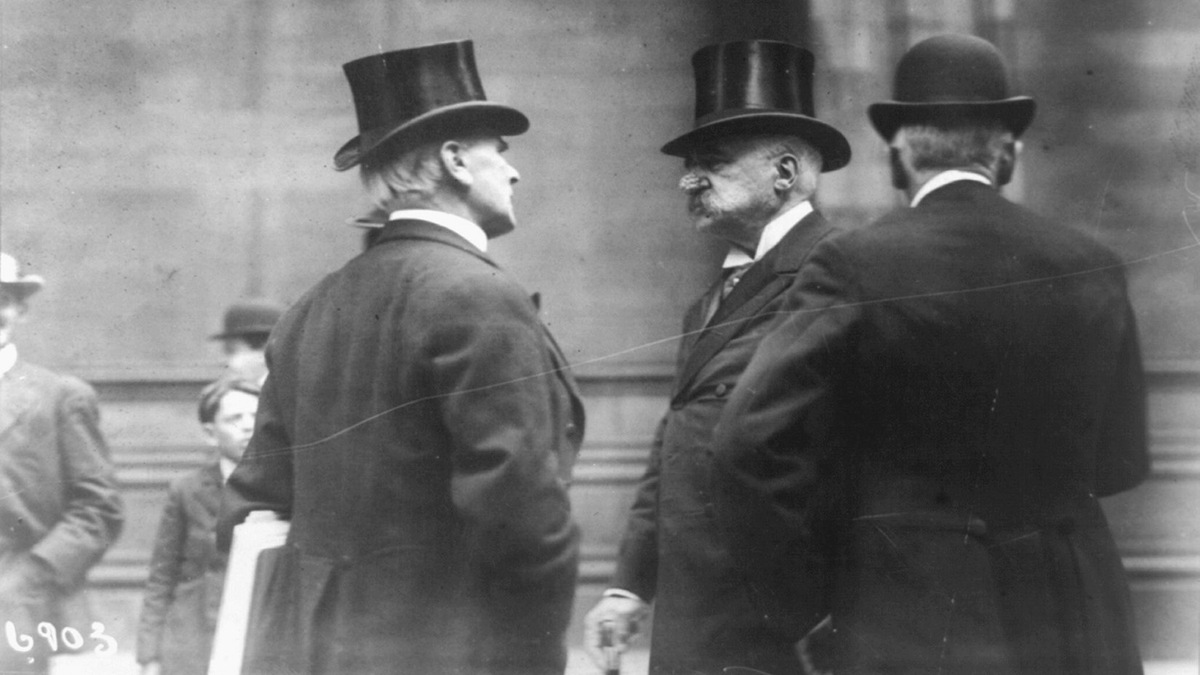 A man in a top hat speaks with another man.