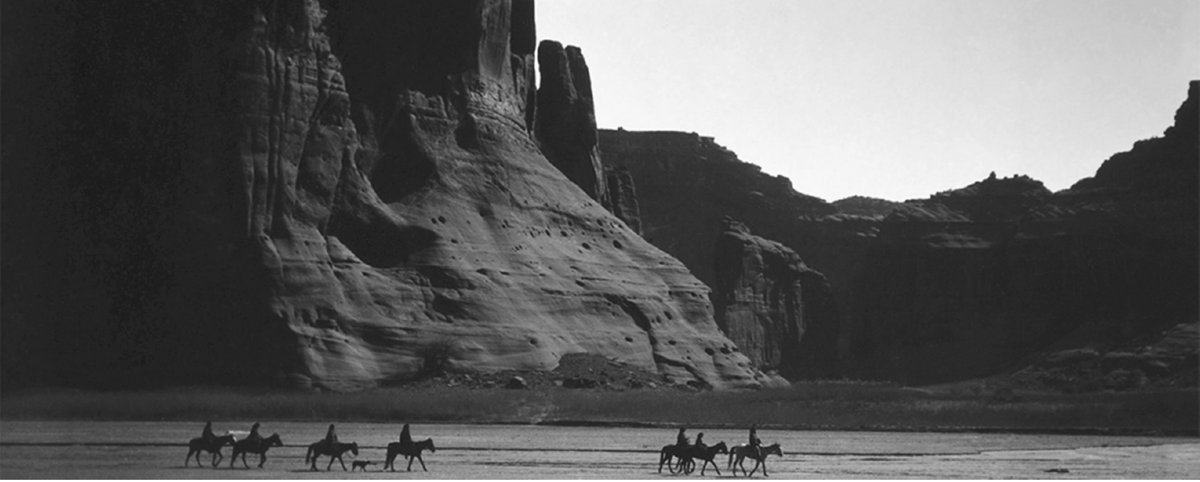 Seven silhouetted riders move through a canyon with tall cliffs behind them.