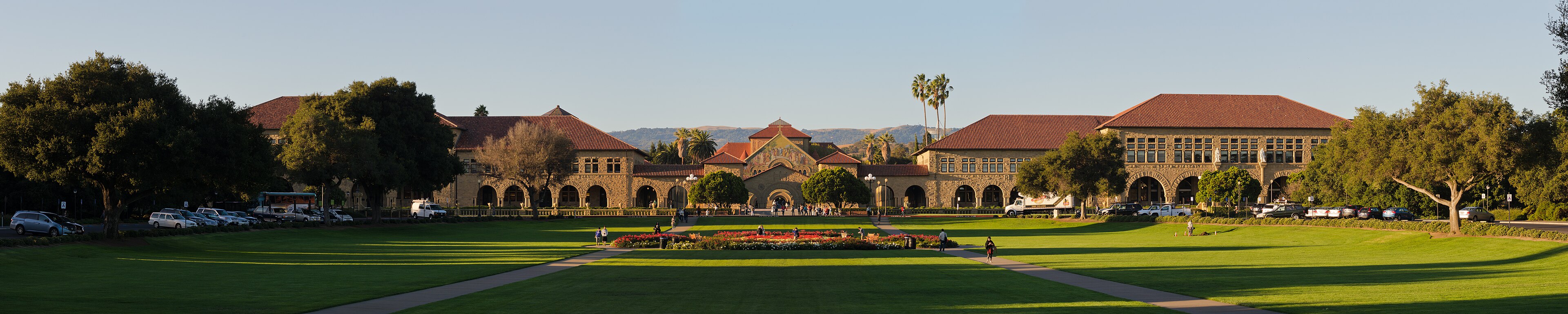 Stanford Main Quad from the Oval, with sunlit sandstone arcades, red-tiled roofs, and an open lawn.